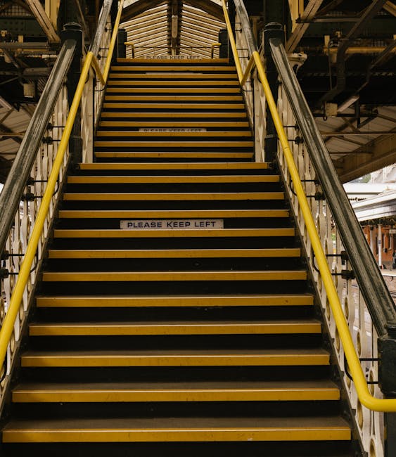 A downward view of outdoor metal stairs with yellow anti-slip strips on each tread, leading from a pavement area to a lower level. The stairs have white-painted metal railings on both sides, with a di