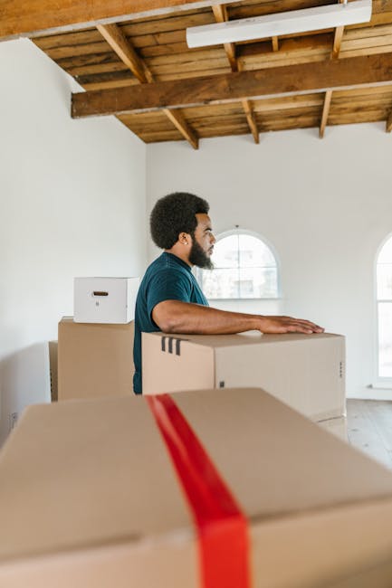 A man with dark curly hair and a beard, wearing a dark teal T-shirt, is standing inside a bright room with white walls and large arched windows, preparing for a home relocation. He is positioned behind and slightly to the right of a stack of cardboard moving boxes, some sealed with red packing tape. The boxes are of various sizes, and one is placed on top of another, with a smaller box to the left. The ceiling features exposed wooden beams and a sloped design, indicating an attic or loft area. Natural light filters through the windows, illuminating the scene. The man appears to be focused on the packing and moving process, part of the furniture transport and moving logistics managed by Removal Company Barnes, as part of their house removals services for the Castelnau moving guide for house removals Barnes SW13, BARNES.