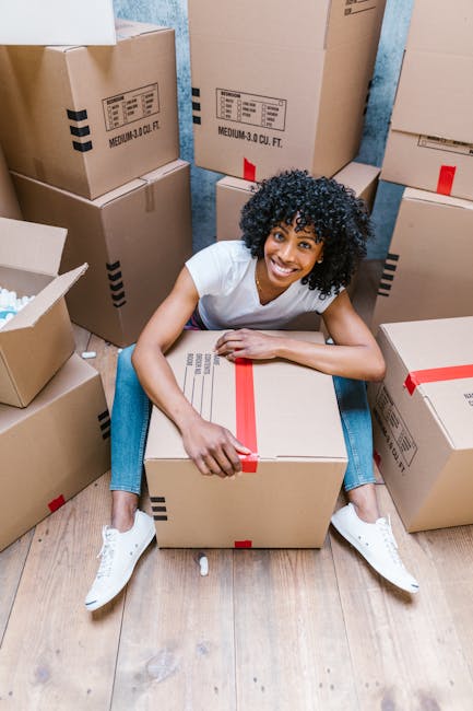 A man and woman are pictured inside a home surrounded by numerous cardboard moving boxes, some of which are open and filled with household items. The man, wearing a white t-shirt and blue jeans, is kneeling on the floor, carefully placing a ceramic plate into a bubble wrap wrap for protection during transportation. The woman, dressed in a light-colored top, stands nearby, looking down at the boxes and packing materials, with her hands resting on her hips. Behind them, there is a large potted plant with broad green leaves, situated against a plain white wall, indicating the early stages of a home relocation process. The scene takes place in a well-lit interior, likely illuminated by natural light, and demonstrates typical packing and loading activities associated with professional removals services provided by Removal Company Barnes, preparing household belongings for safe transport during a house move or furniture transport effort.